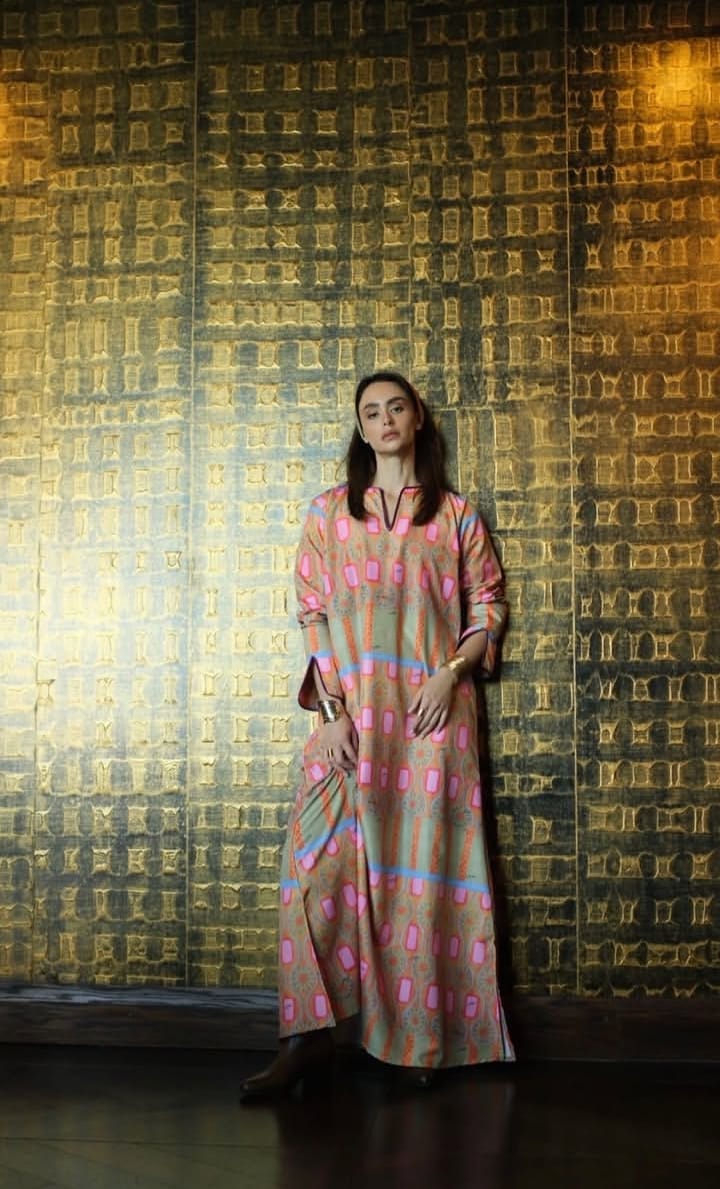 A woman wearing a long-sleeved, knee-length kaftan dress with a pink, orange, and blue geometric pattern, standing in front of a textured gold and black wall.